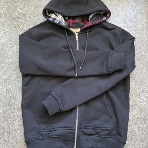 Svart hoodie från Burberry med dragkedja - Svart hoodie från Burberry i bomull med klassiskt rutigt foder i huvan. Tröjan har dragkedja framtill, snörning i huvan och två fickor. Perfekt för dig som gillar stilren design med en touch av lyx. Passar mer i storlek S