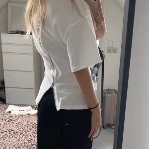 Vit t-shirt med dragkedja - Säljer en vit t-shirt från zara med cool dragkedjedetalj längs hela ryggen. T-shirten har korta ärmar och en något figurnära passform. Perfekt för dig som vill ha en basic med en twist. Materialet känns mjukt och skönt mot huden.