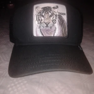 Svart keps Tiger helt ny  - Svart keps från The Farm med broderad tigerpatch framtill. Kepsen har klassisk trucker-modell med mesh baktill och justerbar snapback. Framsidan är i tyg och patchen har detaljer i vitt, beige och svart. Perfekt för dig som vill sticka ut med djurmotiv.
