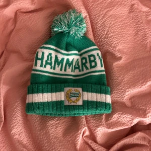 Grön-vit Hammarby mössa med tofs - Snygg grön och vit Hammarby-mössa med tofs på toppen och klubbens logga framtill. Mössan har bred uppvikt kant, stickat mönster med texten 'HAMMARBY' och klassiska ränder. Perfekt för att visa ditt stöd på matchen eller i vardagen. helt oanvänd. nypris 300.