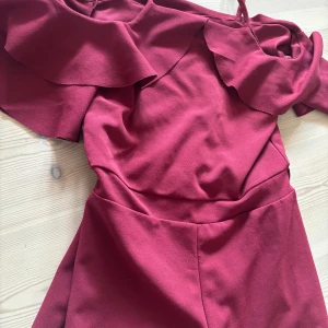 Vinröd kortklänning med volang - Supersnygg vinröd kort byxeress med volangdetaljer upptill och en axelbandslös design på sidan. Dressen har en figurnära passform och är tillverkad i ett mjukt, slätt tyg som känns bekvämt mot huden. Perfekt för dig som vill sticka ut med en färgstark look. Passar perfekt till sommaren och är extremt fin på!!säljer då den är för liten för mig och inte kommmer till användning längre.