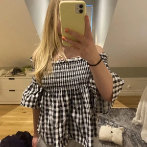 Rutig offshoulder topp från BikBok - Superfin offshoulder topp från BikBok i svartvitt rutigt mönster. Toppen har smockad överdel och vida, korta ärmar som ger en luftig känsla. Perfekt för dig som gillar en söt och trendig stil. Gjord i mjuk bomull som känns skön mot huden.