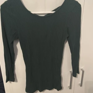 Mörkgrön ribbad topp från AMISU - Mörkgrön långärmad topp från AMISU i ribbad bomullsmix, köpt inne på H&M. Snygg detalj med korsade band i ryggen och figurnära passform. Perfekt till jeans eller kjol för en trendig look💕