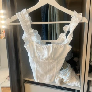 Vit volanglinnetopp  - Supersöt vit linnetopp med breda volangprydda axelband och lätt rynkad struktur. Toppen är croppad och har en fyrkantig halsringning som ger en trendig vibe. Perfekt för varma dagar och enkel att styla med jeans eller kjol.