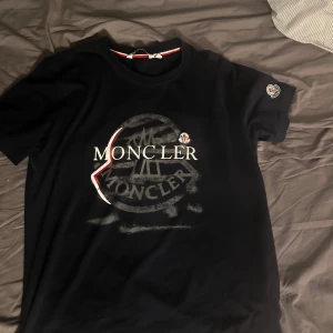 Svart Moncler t-shirt med logga - Snygg svart t-shirt från Moncler med stor tryckt logga framtill och broderad patch på ärmen. Klassisk rund halsringning med röd och vit detalj på insidan av kragen. Perfekt för dig som gillar streetwear och exklusiva märken. Storlek L men passar M