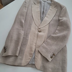Beige kavaj från Emporio Armani - Snygg beige kavaj från Emporio Armani i linne med breda slag och dekorativa knappar vid ärmslutet. Kavajen har en ljusare beige kontrast på slaget och klassisk passform. Perfekt för dig som vill ha en stilren och exklusiv look. Inga fläckar. 