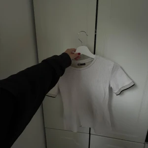 Vit ribbad t-shirt från Zara - Vit ribbad t-shirt från Zara med korta ärmar. Ärmsluten har en snygg detalj med svarta och guldiga ränder som ger en cool kontrast. T-shirten är i ett stretchigt och mjukt material som sitter skönt på kroppen.