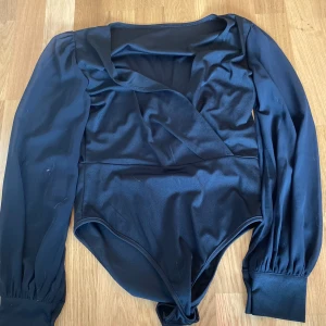 Svart omlott bodysuit med långa ärmar - Snygg svart bodysuit med omlott-skärning och långa puffärmar. Djup v-ringning och breda muddar vid ärmsluten ger en trendig look. Perfekt att styla med jeans eller kjol för en cool outfit.