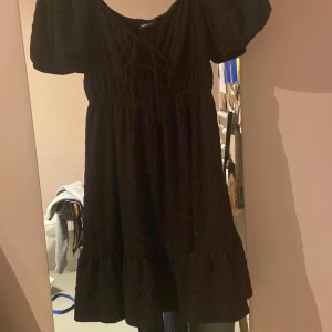 Svart kortklänning från Gina Tricot - Supersöt svart kortklänning från Gina Tricot med puffiga korta ärmar och snörning framtill. Klänningen har en luftig passform och volang nertill, perfekt för en trendig look. Tillverkad i mjukt tyg som känns skönt mot huden.