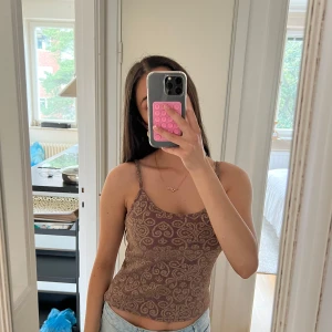 Brun mönstrad linnetopp med smala band - Säljer en brun/lilla linnetopp med smala axelband och ett unikt paisley-liknande mönster i beige. Toppen är croppad och sitter tajt mot kroppen, perfekt till jeans eller shorts. Materialet känns mjukt och stretchigt, vilket gör den superbekväm och snygg till sommaren.