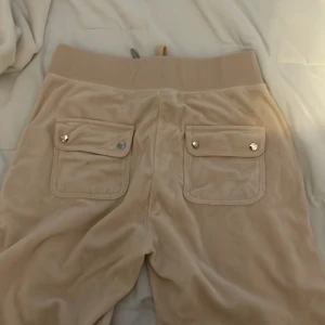 Beiga mjukisbyxor Juicy Couture, Storlek: S - Supermjuka beiga mjukisbyxor från Juicy Couture med bred resår i midjan och snörning framtill, denna byxan har endast använts 1 gång pga den var för liten för just mig😊. Byxorna är flared, stora fickor med lock och tryckknappar bak samt sidofickor fram. Klassisk Juicy-logga broderad på ena fickan.