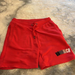 Röda shorts från Hoodrich XS - Röda shorts från Hoodrich med broderad logga i svart, grått och vitt på ena benet. Modellen har snörning i midjan och sidofickor. Mjukt material som känns skönt mot huden, perfekt för chill dagar eller träning.