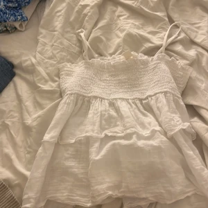 Vit volangig linnetopp Gina Tricot - Supersöt vit linnetopp från Gina Tricot med smala axelband, smockad överdel och dubbla volanger nertill. Tunt och luftigt tyg som känns perfekt för varma dagar. Snyggt rynkad upptill och lätt ruffig kant för extra detalj.