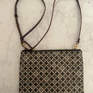 Axelväska från By Malene Birger, svart/beige - Snygg axelväska från By Malene Birger med justerbar rem och dragkedja. Väskan har ett grafiskt mönster i svart och beige, guldiga metalldetaljer och är tillverkad i tåligt syntetmaterial. Perfekt för dig som gillar stilrena accessoarer med en twist.