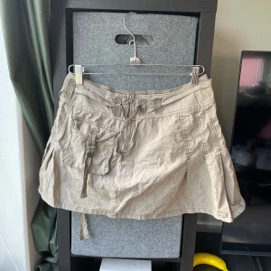 Beige utilitykjol från BDG Urban Outfitters - Aldrig använd!! Säljer en beige utility-inspirerad kortkjol från BDG Urban Outfitters. Kjolen har stora fickor med spännen, bälteshällor och dragkedja framtill. Perfekt för dig som gillar streetstyle och praktiska detaljer.