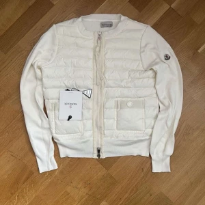 Vit cardigan jacka från Moncler - Säljer denna exklusiva Moncler cardigan/jacka i krämvitt – ett måste i varje garderob. Dunvadderad front och stickade ärmar i bomull ger både stil och komfort. Äkta vara med Moncler-logga på ärmen, originaltaggar samt digitalt kvitto som bevis på köp. Prissänkning kan diskuteras endast om du är seriös 