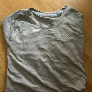 Grå mönstrad t-shirt från Gymshark - Snygg grå t-shirt från Gymshark med diskret geometriskt mönster och logga på bröstet. Perfekt för träning med sitt lätta och stretchiga material. Klassisk rund halsringning och korta ärmar för en sportig look.