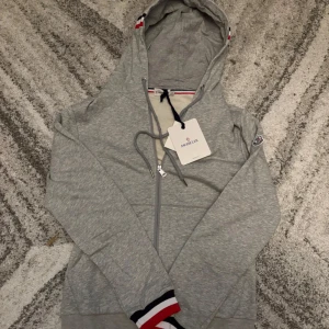 Grå Moncler kofta - Snygg grå hoodie från Moncler med hel dragkedja, huva och snörning. Tröjan har långa ärmar och Moncler-logga på ärmen. Ribbade muddar med röd, vit och marinblå rand ger en sportig touch. Perfekt för dig som gillar stilren streetwear.