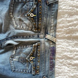 Low waist bootcut jeans - Blå bootcut jeans från Plastik by Sly med snygga nitar och broderier på bakfickorna. Låg midja, raka ben med lätt utsvängning och coola detaljer som kontrastsömmar och broderad logga i lila. Perfekta för dig som gillar statement-jeans med edge.