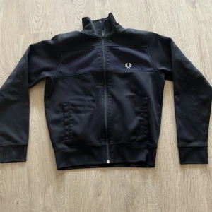 Svart track jacket från Fred Perry - Svart track jacket från Fred Perry med klassisk logga broderad på bröstet. Jackan har hög krage, hel dragkedja framtill och två fickor. Tillverkad i mjukt syntetmaterial som ger en sportig och clean look. Perfekt för dig som gillar streetwear och stilren design.