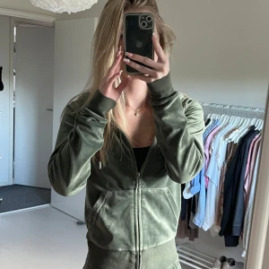 Olivgrön velourjacka med dragkedja - Snygg olivgrön zip-up jacka i mjuk velour med huva och fickor framtill. Jackan har långa ärmar, bred mudd i midjan och är perfekt för en chill och trendig look. Passar dig som gillar sportig och avslappnad stil. + Byxor