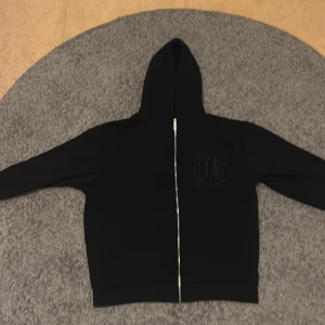 Svart hoodie med dragkedja och tryck - Svart hoodie med hel dragkedja och huva. På bröstet finns ett stort tryck i form av en gotisk bokstav. Tröjan har långa ärmar och en clean look, perfekt för dig som gillar streetwear. Materialet känns mjukt och bekvämt, troligen bomull eller polyester.