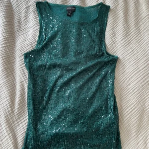 Grön paljettopp från River Island - Säljer en glittrig grön ärmlös topp från River Island. Toppen är täckt av små paljetter som ger ett riktigt snyggt skimmer. Modellen är rak och har breda axelband, perfekt för dig som vill sticka ut lite extra.