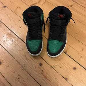Nike Air Jordan 1 svart/grön sneakers - Säljer ett par Nike Air Jordan 1 sneakers i svart och grönt skinn med vita sulor. Klassisk high-top modell med svart snörning och röda Nike-detaljer på plösen. Perfekta för dig som vill sticka ut med en ikonisk och sportig look.
