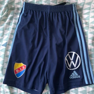 Blå DIF fotbollsshorts från Adidas - Snygga marinblå fotbollsshorts från Adidas med DIF-emblem och Volkswagen-logga. Shortsen har ljusblå ränder längs sidorna och resår i midjan. Perfekta för fotbollsträning eller match. Materialet är lätt och andas, vilket gör dem bekväma att röra sig i. Endas använda några gånger, ny pris 329. Priset kan diskuteras vid snabb affär 