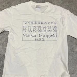 Maison Margiela vit t-shirt med tryck - Vit t-shirt från Maison Margiela med ikoniskt siffertryck och logga på bröstet. Klassisk rund halsringning och korta ärmar. Tillverkad i mjuk bomull som känns skön mot huden. Perfekt för dig som gillar stilrena och trendiga plagg med designer-touch. Lägger ut igen för att inte fick den såld!!!!!!