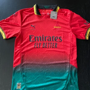 AC Milan x Off White x Puma fotbollströja L - AC Milan 2025 limited edition fotbollströja från samarbetet mellan Off White och Puma. Röd upptill med grön nedre del i snygg gradient, svarta och gula detaljer på krage och ärmslut. Broderade loggor, 'Emirates Fly Better' tryck och tillverkad i lätt funktionsmaterial.