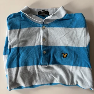 Randig pikétröja från Lyle & Scott - Snygg pikétröja från Lyle & Scott med breda blå och vita ränder. Klassisk krage, tre knappar framtill och den ikoniska gula loggan broderad på bröstet. Perfekt för en avslappnad och sportig stil.