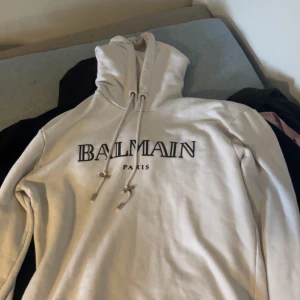 Vit hoodie från Balmain Paris - Säljer en clean vit hoodie från Balmain Paris med stor logga framtill. Tröjan har huva med snörning och är tillverkad i mjukt material som känns skönt mot huden. Perfekt för dig som gillar exklusiva streetwear-vibes och vill sticka ut.