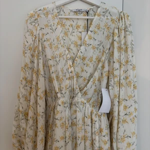 Blommig långklänning från NA-KD - Superfin långklänning från NA-KD i vit med gula blommor. Klänningen har v-ringning, långa puffärmar och omlott-look framtill. Tillverkad i ett lätt, plisserat material som ger en flowy känsla. Perfekt för sommarens alla tillfällen! Aldrig använd.