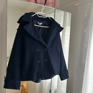 Mörkblå kappa med dubbelknäppning - Superfin, stilren kappa i kortare modell. Märkt som M/L men passar även dig som vanligtvis har S/M för en mer oversized look. Använd men är i ett fint skick!! Säljer då den inte kommer till användning längre