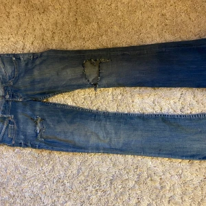 Blå jeans med hål på från H&M - Säljer ett par blå mammajeans från H&M med snygga slitningar på benen. Jeansen har klassisk femficksdesign, rak passform och är tillverkade i mjukt bomullsjeans. Perfekta för en avslappnad och trendig look. Har en liten slitning på sidan