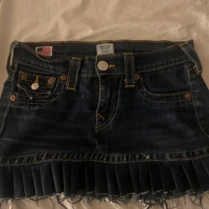 True Religion jeanskjol med volangkant - Mörkblå jeanskjol från True Religion med klassiska kontrastsömmar, fem fickor och snygg volangkant nertill. Kjolen har rå fransad nederkant och detaljerade bakfickor med knappar. Oanvänd 