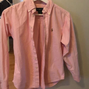 Rosa skjorta från Ralph Lauren - Klassisk rosa skjorta från Ralph Lauren med button-down krage och broderad logga på bröstet. Skjortan har långa ärmar och knäppning framtill. Perfekt för en stilren och avslappnad look.finns en liten fläck fast samma färg som skjortan så det syns inte mycket. Tror dock att det går bort i tvätten och jag tvättar givetvis den innan jag skickar . 
