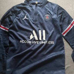 PSG långärmad fotbollströja blå - Snygg långärmad PSG-tröja i mörkblått med vita och röda detaljer på ärmarna. Tröjan har halv dragkedja, Paris Saint-Germain-logga på bröstet och Jordan-logga. Materialet är lätt och andas, perfekt för fotbollsträning eller att bära till vardags.