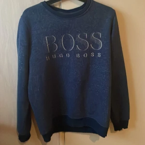Blå sweatshirt från Hugo Boss - Snygg blå sweatshirt från Hugo Boss med stor logga framtill. Tröjan har rund halsringning, långa ärmar och ribbade muddar vid ärmslut och nederkant. Perfekt för en chill och stilren look.