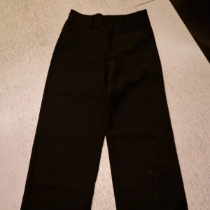 Svarta kostymbyxor XOXO, strl 152 - Stilrena svarta kostymbyxor från XOXO, storlek 152. Klassisk rak modell med bälteshällor och knapp framtill. Perfekta för dig som vill ha en clean och enkel look. Materialet känns mjukt och har en lätt glans.