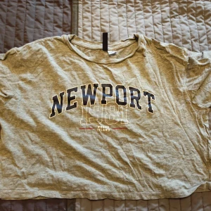 Grå t-shirt Newport 1981 Divided XS - Grå t-shirt från Divided med tryck 'NEWPORT 1981 Rhode Island' i svart och vitt framtill. Klassisk rund halsringning och korta ärmar. Skön och avslappnad passform, perfekt till jeans eller shorts. Oversized kort magtröja