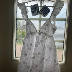 Vit broderad kortklänning med volang - Superfin vit kortklänning med broderade blommor, volangdetaljer på axelbanden och öppen rygg med knyt. Aldrig använd, endast testad! Köpt utomlands. Köpt för 745 kr! 