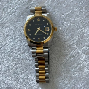 Reginald klocka i guld och silver - Snygg Reginald klocka med svart urtavla, gulddetaljer och datumvisning. Armbandet är i tvåtonad metall med både guld- och silverfärgade länkar. Klockan har romerska siffror och glittriga indexmarkeringar, samt en klassisk rund boett och guldfärgad krona. Nya klockan med nya batteri, 389kr/st 