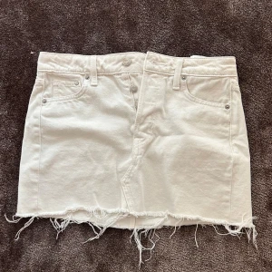 Vit jeanskjol från &Denim XS - Vit kort jeanskjol från &Denim i storlek XS. Kjolen har råa, fransiga kanter nertill och klassiska jeansdetaljer som fickor fram och bak samt bälteshällor. Snygg asymmetrisk design med dubbla knappar framtill för en unik look.