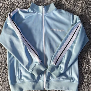 Ljusblå track jacket från Palm Angels - Säljer nu min ljusblåa palm angels tröja då jag inte tycker om den längre. Den är använd flertal gånger så den är lite sliten. Den är storlek S men passar även M väldigt bra. Den har en liten fläck på sidan av armen. Dragkedjan är också lite krånglig ibland. Har ni nån fråga är det bara o skriva 
