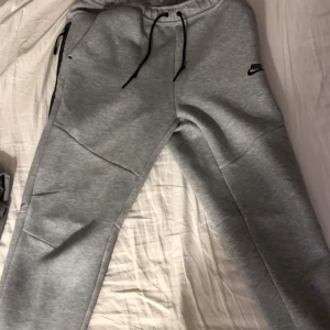 Grå mjukisbyxor från Nike, storlek L - Snygga grå mjukisbyxor från Nike med svarta dragkedjor på fickorna och justerbar snörning i midjan. Byxorna har en modern passform och är tillverkade i mjukt bomullsmaterial. Perfekta för chill eller träning, med diskret Nike-logga på benet.