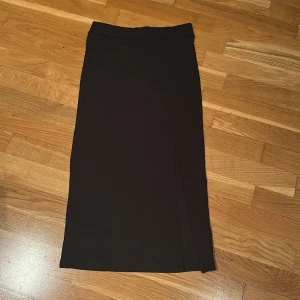 Svart ribbad kjol från Gina Tricot - Snygg svart ribbad kjol från Gina Tricot i en smickrande slim passform. Kjolen har en diskret slits som ger en extra touch. Perfekt för att styla med olika toppar och accessoarer.