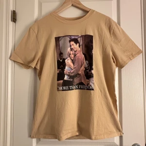Beige t-shirt med Friends-tryck från NA-KD - Säljer en beige t-shirt från NA-KD med tryck från TV-serien Friends. Framsidan har ett stort motiv av karaktärerna Rachel och Ross med texten 'More Than Friends'. Baksidan visar flera scener från serien. Perfekt för fans av serien!