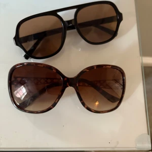 Två par solglasögon - Säljer två par solglasögon. Ett par med stora, runda bågar i leopardmönster och guldiga detaljer på sidorna. Det andra paret har en mer klassisk, fyrkantig form i svart med bruna glas. 50 kr/solglasögon